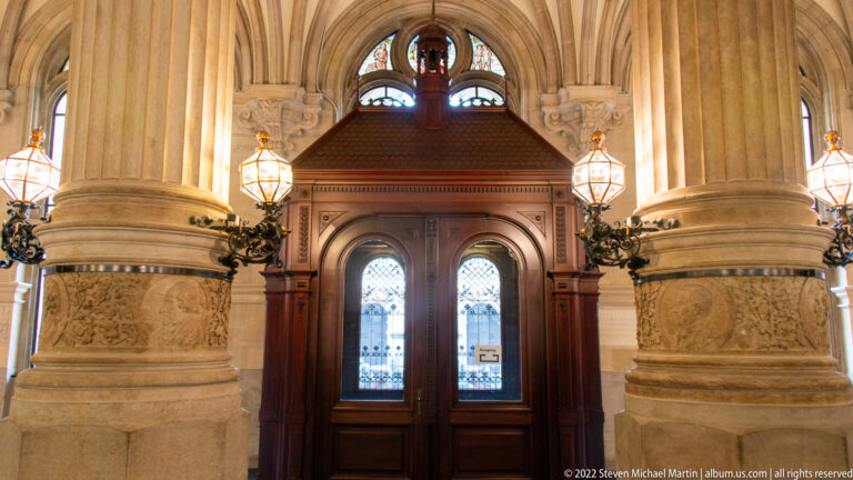 Dark wooden double doors with leaded glass framed by massive carved stone columns and ornamental lamps inside Hamburg Rathaus.