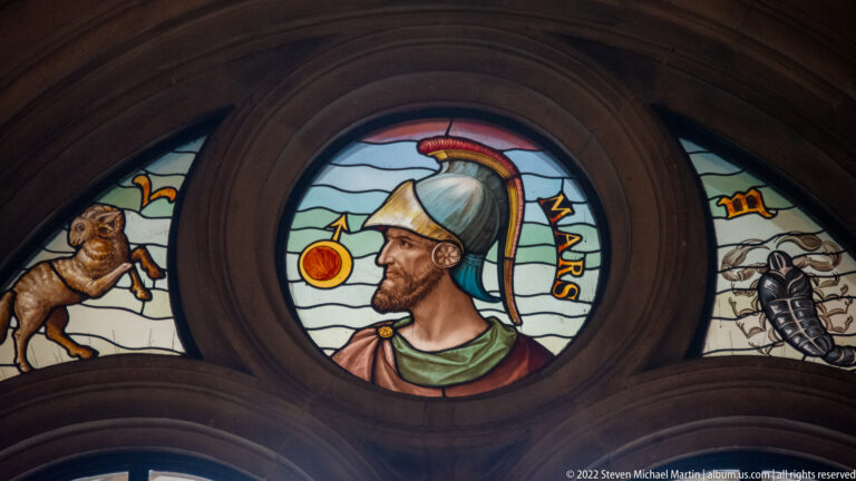 Stained-glass medallion of a helmeted male figure labeled “MARS,” surrounded by smaller symbolic panels.