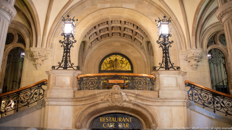 Ornate Rathaus stair landing with twin lamps, wrought-iron railings, stained glass, and a restaurant entrance below.