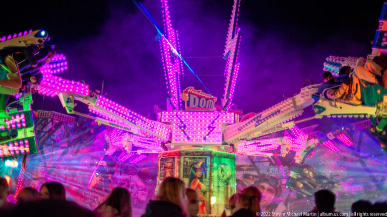 Nighttime amusement ride covered in magenta and green lights, with spectators in front and a small Dom sign above.