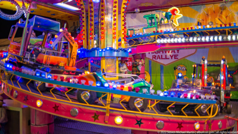 Small truck-shaped children's ride vehicle on a brightly lit platform with colorful fairground scenery behind it.