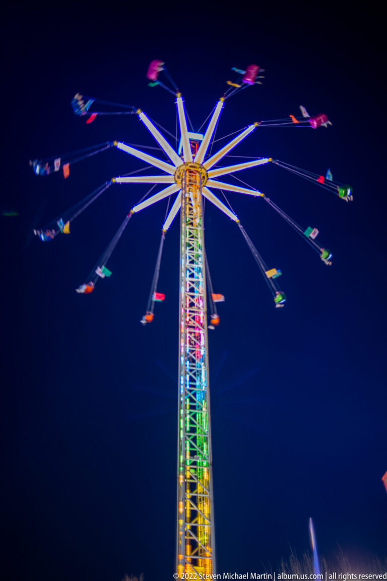 Upward view of a multicolored tower swing ride with long arms and blurred suspended chairs against the night sky.