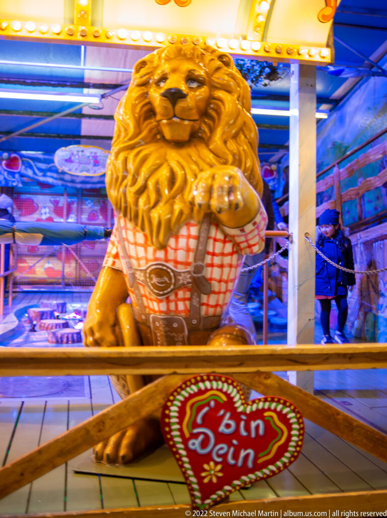 Golden lion figure holding a red-and-white checked basket above a heart-shaped sign amid blue fairground lights.