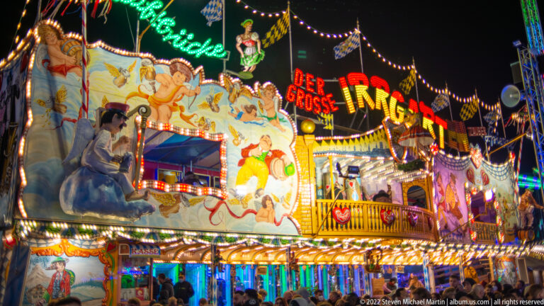 Brightly illuminated Der Grosse Irrgarten funhouse façade with painted figures, flags, mirrors, and visitors below.