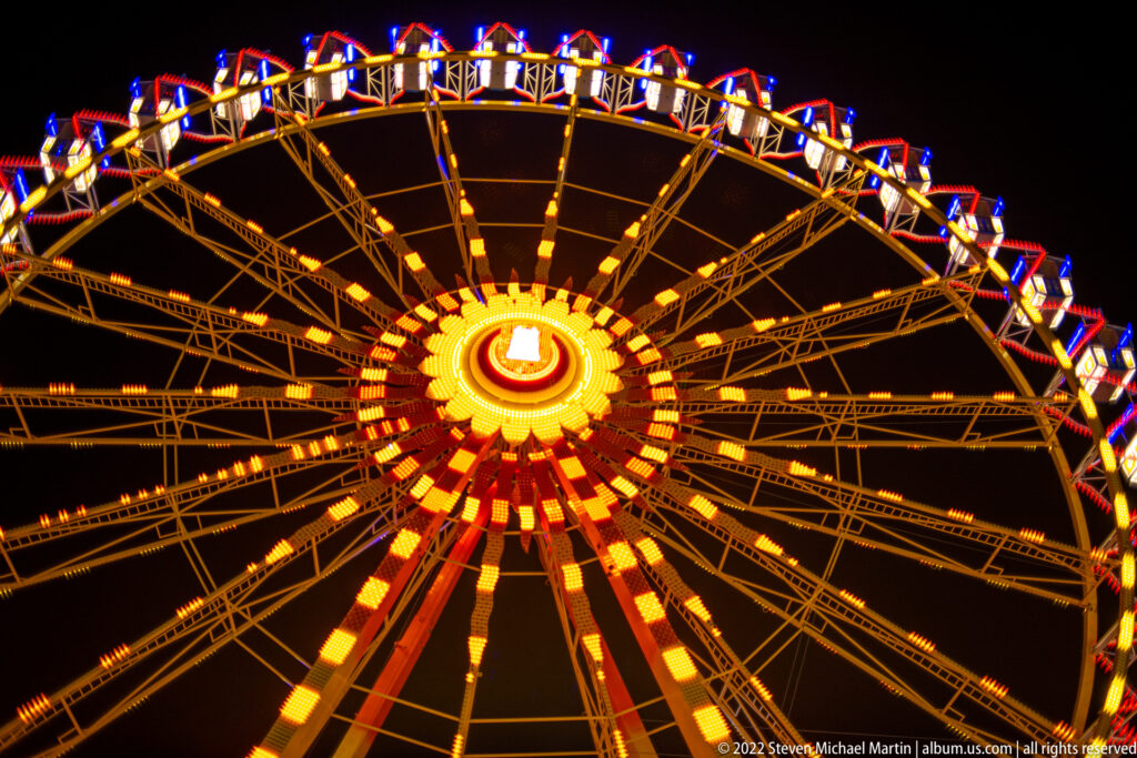 Low-angle view of a Ferris wheel with glowing amber spokes and a bright circular hub against a black night sky.
