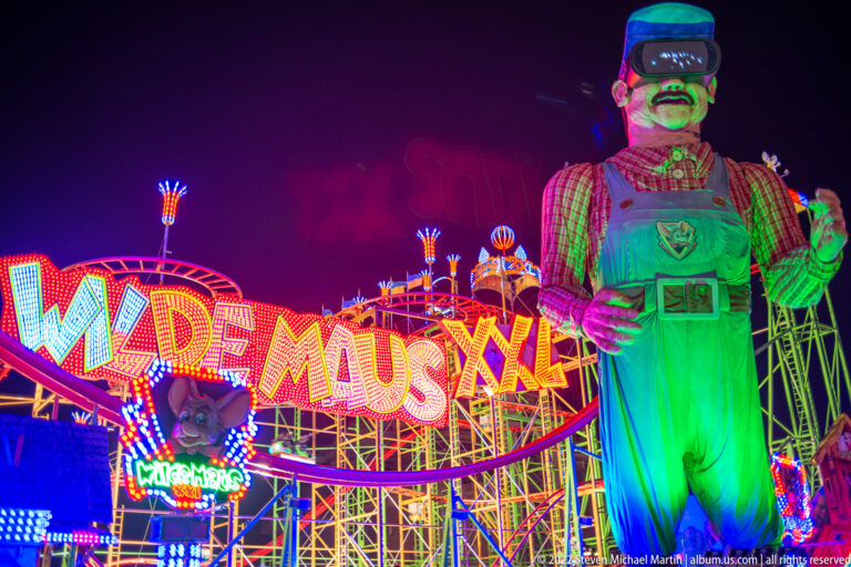 Giant green mouse mascot in lederhosen beside the illuminated Wilde Maus XL roller coaster sign and steel track.