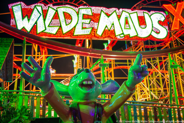 Green cartoon mouse mascot with raised arms beneath the Wilde Maus sign and orange roller-coaster supports.