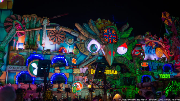 Haunted fairground façade filled with glowing monster faces, masks, dark-fantasy props, and silhouettes of visitors below.