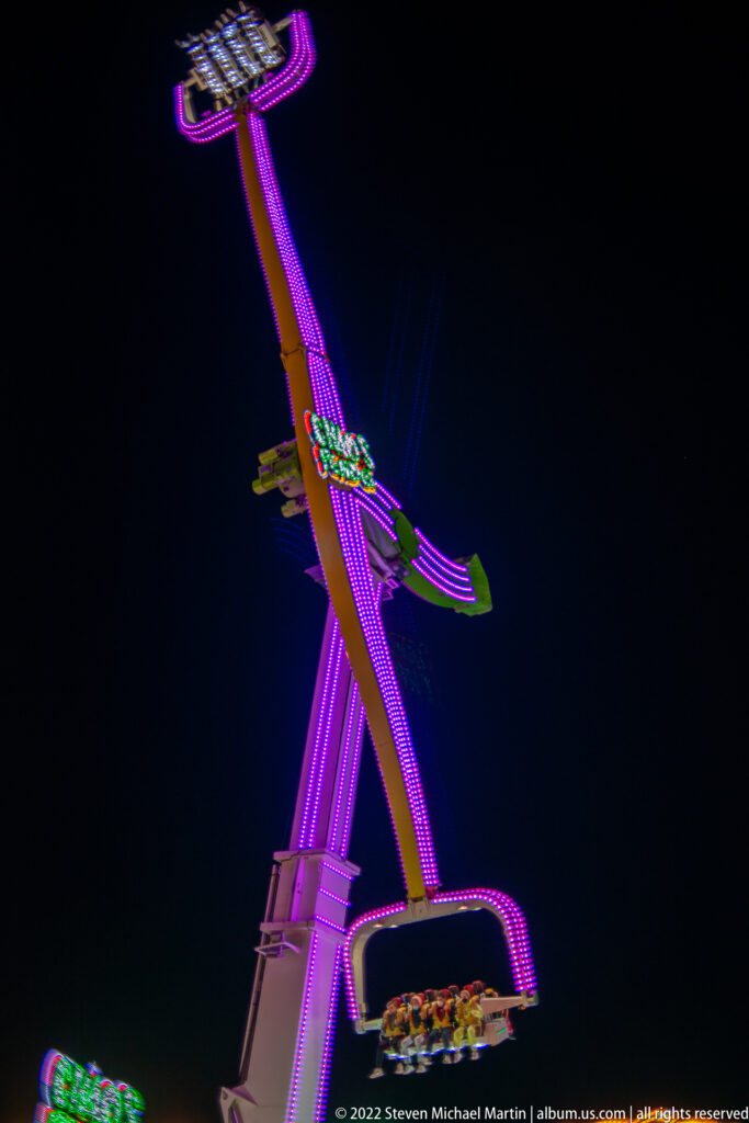 Purple-lit thrill ride with a long angled arm and gondola moving high against the black night sky.