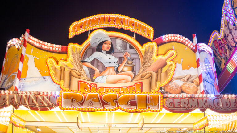 Illuminated Schmalzkuchen food stall with a seated female figure, wing-like decoration, and large red lettering.