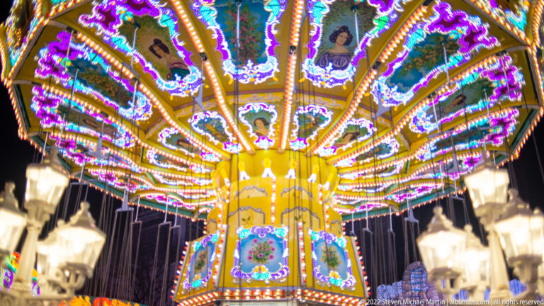 Upward view beneath a chair-swing carousel canopy with radiating lights, painted panels, and hanging chains.