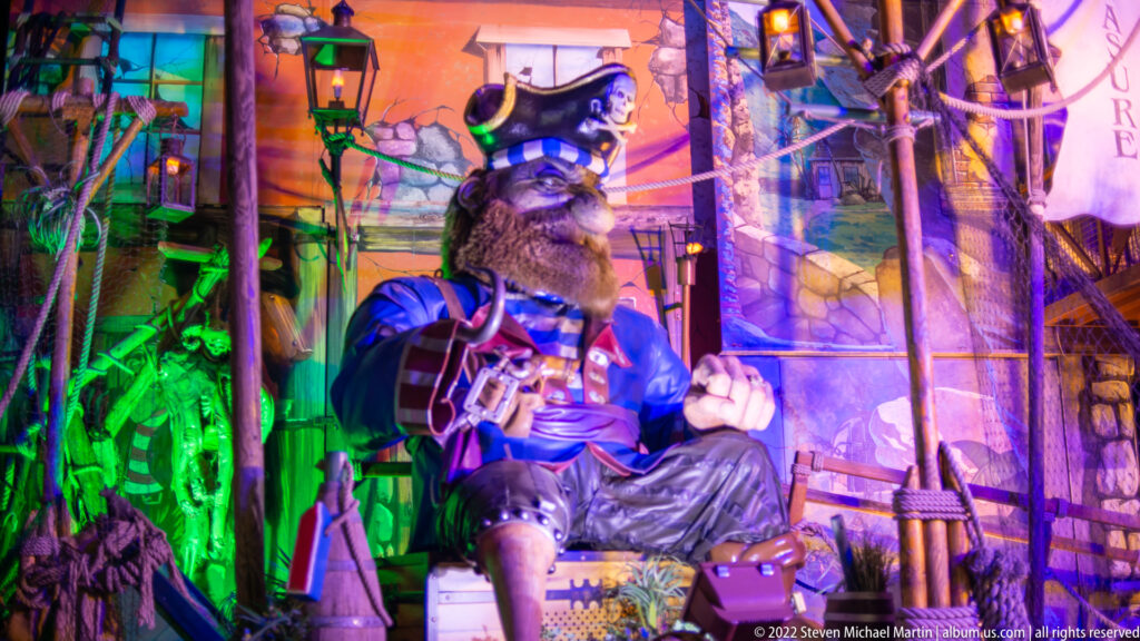 Bearded pirate figure in blue coat and tricorn hat posed among ropes and nautical scenery under colored lights.