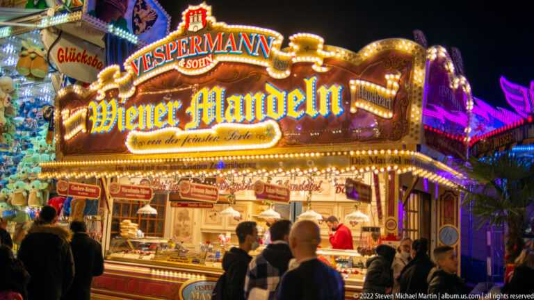 Crowded Vespermann Wiener Mandeln stall with ornate illuminated lettering, rows of bulbs, and customers at the counter.