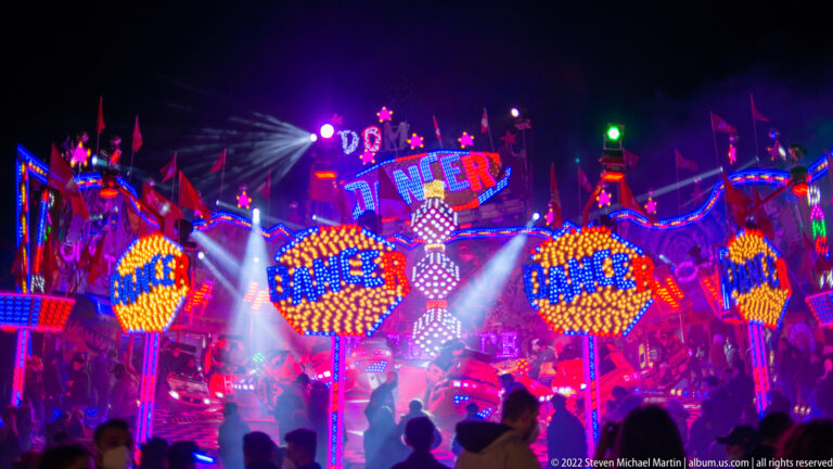 Crowd silhouettes in front of an ENTER-branded thrill ride glowing with red, blue, and amber lights and overhead spotlights.