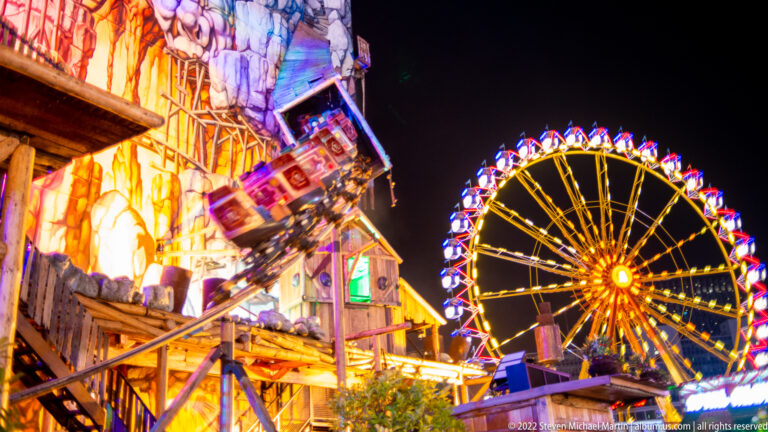 Illuminated Ferris wheel rising behind a colorful angular fairground attraction at night.