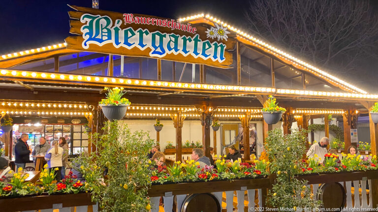 Bauernschänke Biergarten pavilion with bulb-lined gable, flower boxes, greenery, and diners beneath the sign.
