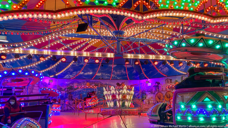 Circular amusement ride with cars beneath a richly decorated canopy covered in multicolored lights.
