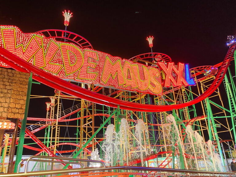 Wilde Maus XL sign following a red roller coaster track above green supports and illuminated fountain jets.