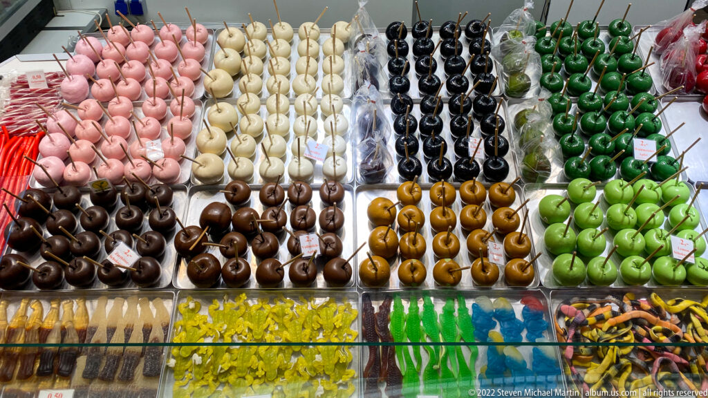 Neat rows of candy-coated apples in pink, white, brown, black, green, and caramel above assorted sweets.
