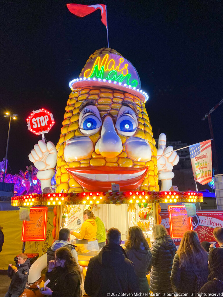 Giant smiling corn mascot above a Mais food stall, with customers below and a red STOP sign at left.