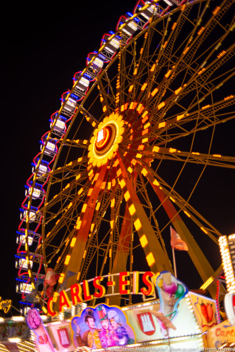 Ferris wheel glowing above a colorful Carls Eis stand at night, with orange spokes and illuminated rim lights.