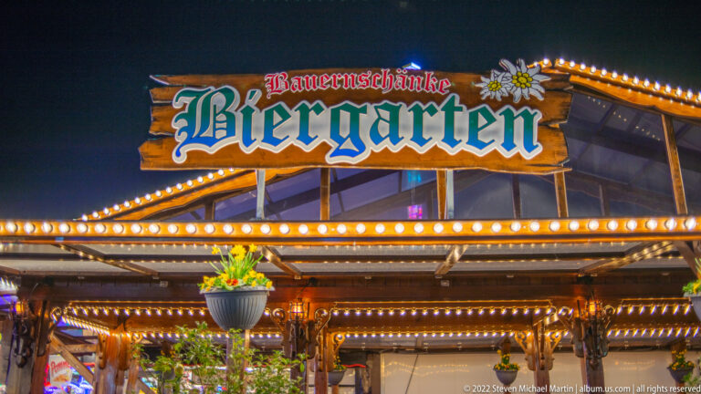 Bauernschänke Biergarten sign above a timber-style fairground pavilion with warm bulbs and hanging greenery.