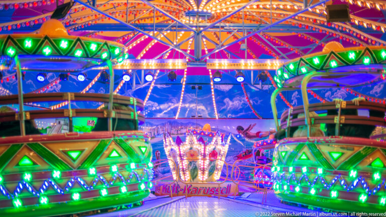 Green children's carousel vehicles beneath a multicolored canopy, edged with blue and green lights.