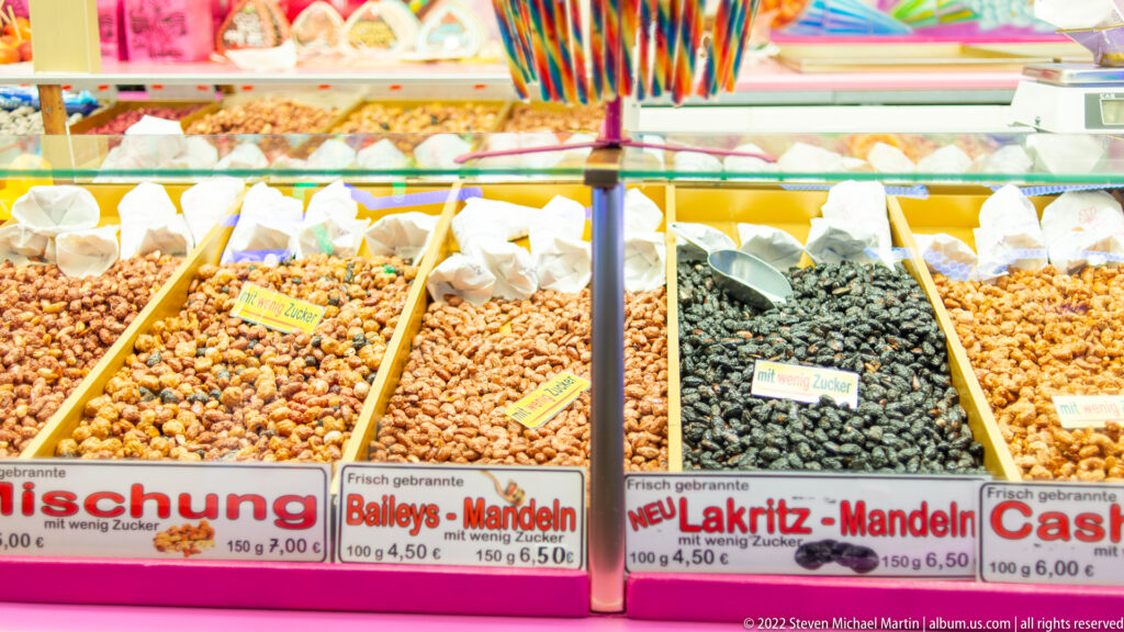 Rows of open trays filled with roasted and coated nuts, including labeled Baileys-Mandeln and Lakritz-Mandeln varieties.