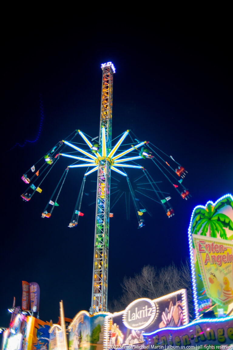 Tall illuminated swing ride with chairs spinning outward high above fairground signs at night.