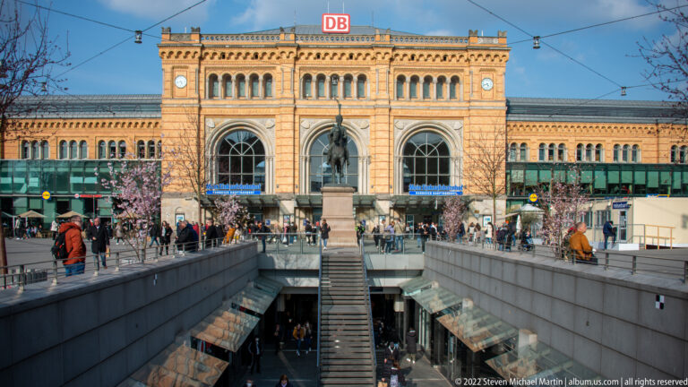 Hannover Hauptbahnhof façade behind stairs to an underground passage, with pedestrians, tram wires, and the red DB logo.