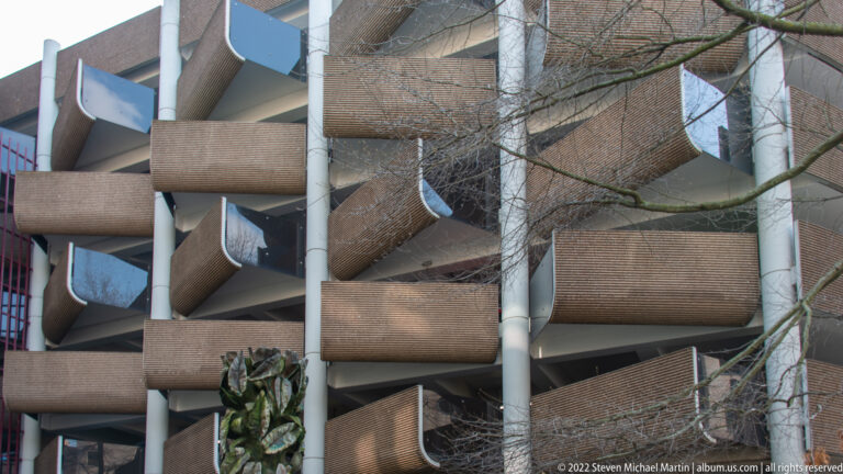 Close view of stacked projecting balconies on a modern brick-and-concrete building, partly screened by bare branches.