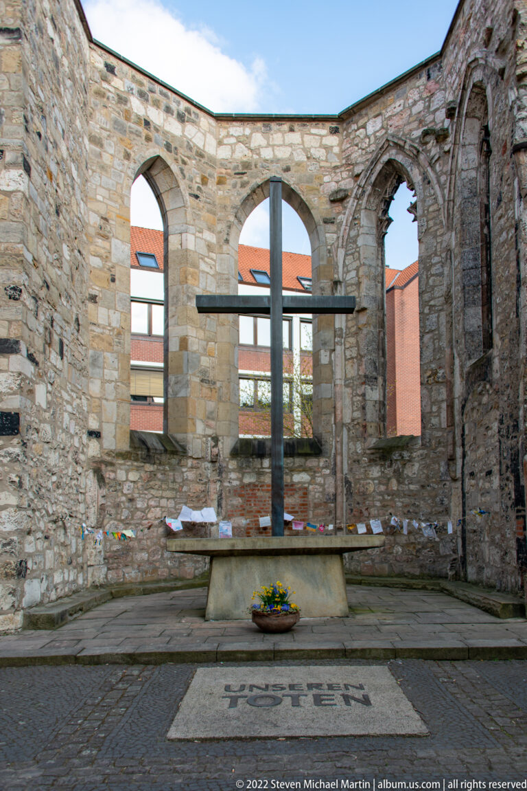 Large cross inside the roofless Aegidienkirche ruins, framed by tall arched window openings and weathered masonry.
