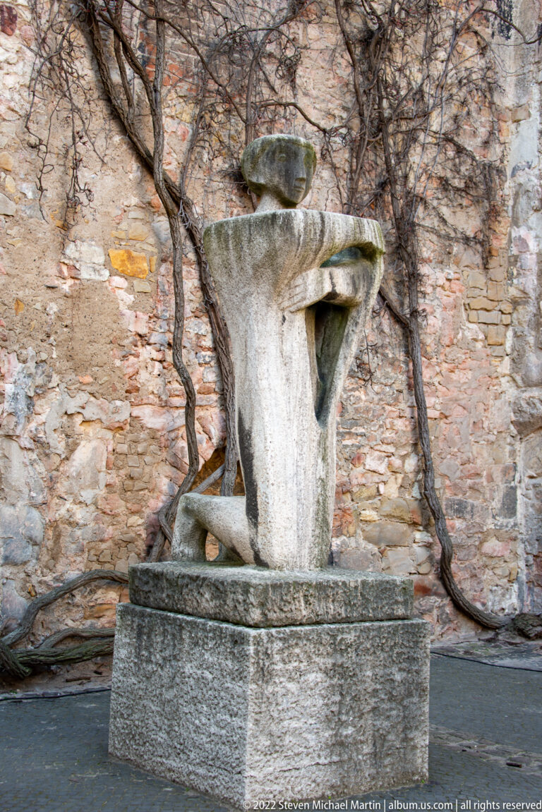 Abstract pale stone sculpture on a plinth against the vine-covered interior wall of Aegidienkirche.