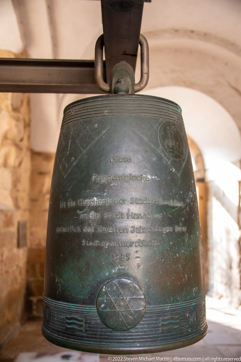 Dark bronze Hiroshima Peace Bell hanging from a timber support inside Aegidienkirche, with German inscription on its surface.