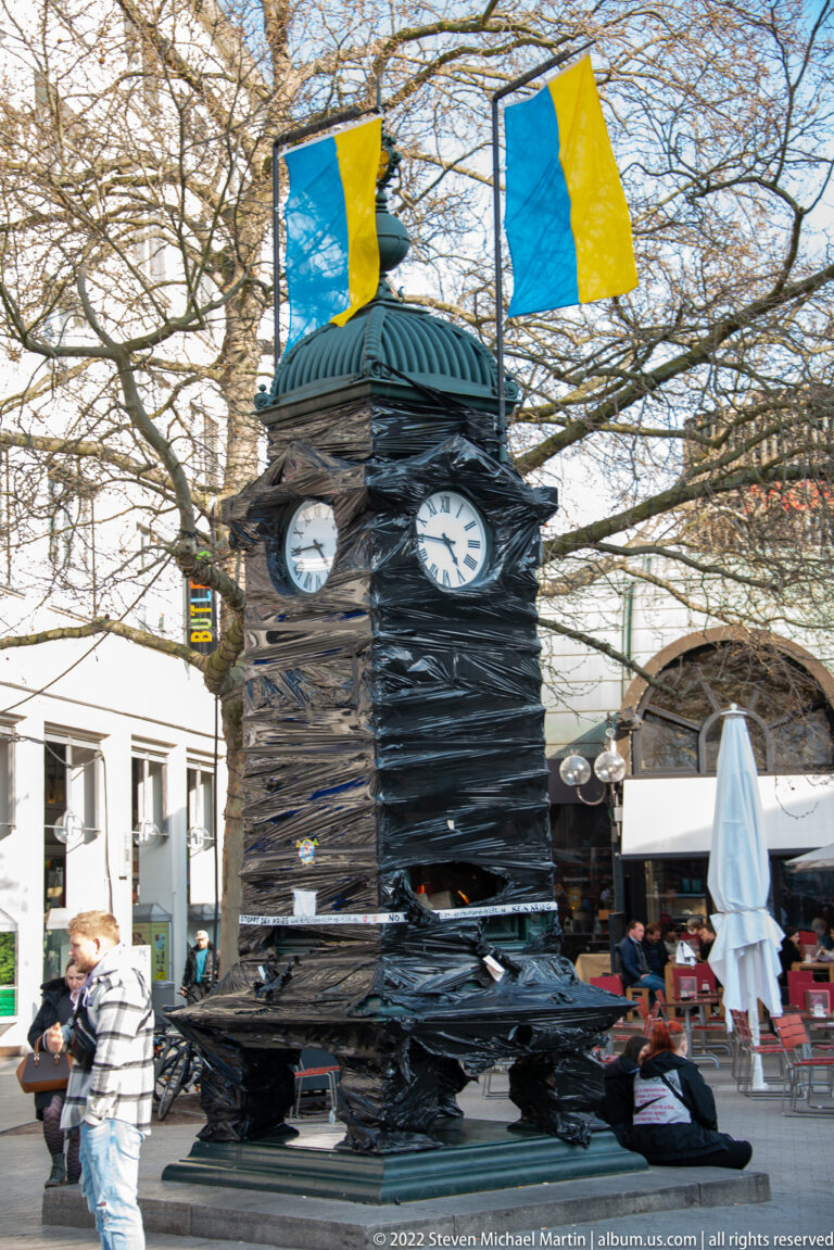 Ornate black Kröpcke street clock with two clock faces beneath blue-and-yellow flags and leafless trees.