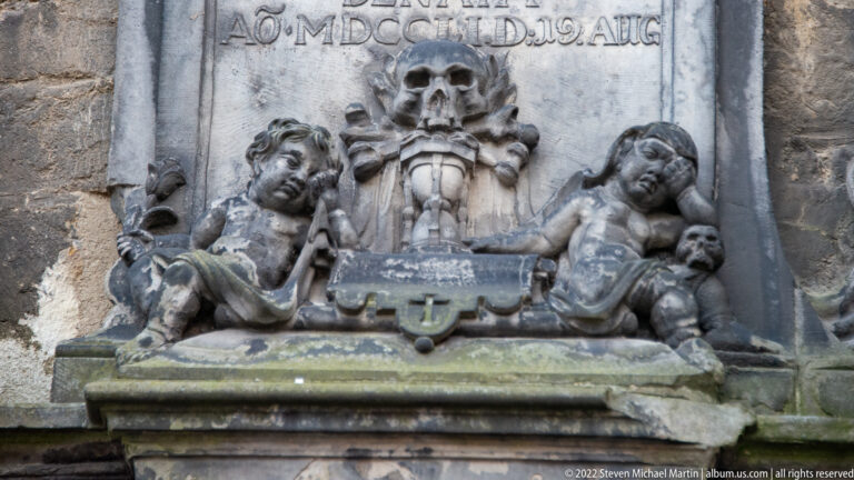 Weathered stone funerary relief with central skull, inscribed panel, and reclining mourning figures.