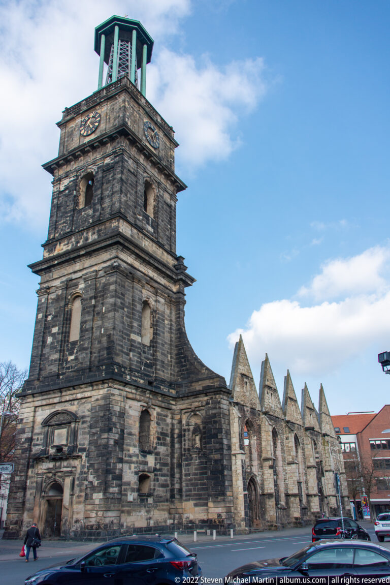 Street view of Aegidienkirche with dark tower, roofless gabled walls, and cars passing in front.
