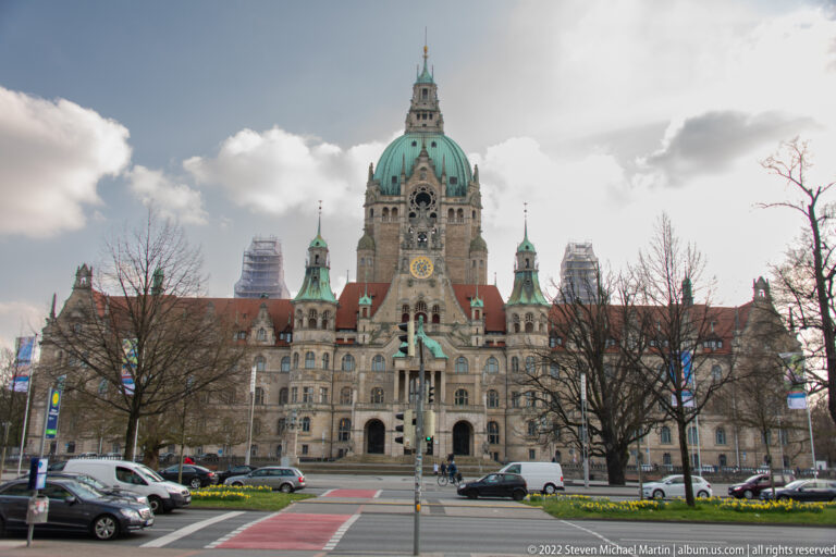 Frontal distant view of Hannover’s Neues Rathaus with large green dome, clock, red roofs, and smaller towers.