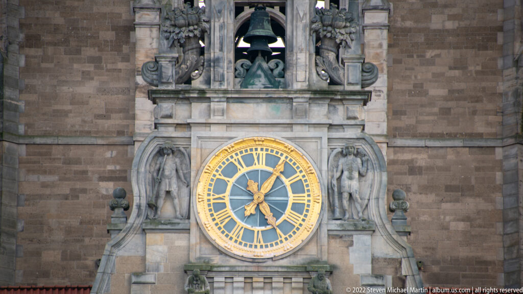 Large blue-and-gold clock on the Neues Rathaus, flanked by stone figures with a bell opening above.