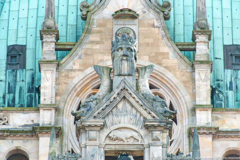 Large bearded stone head above a triangular pediment on the Neues Rathaus, framed by copper roof panels.