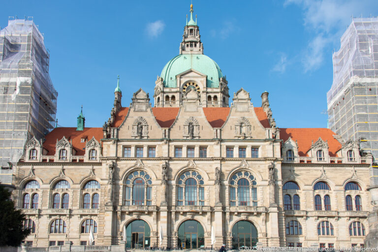 Side view of the Neues Rathaus with green dome, red roofs, three large arched windows, and scaffolding at both edges.