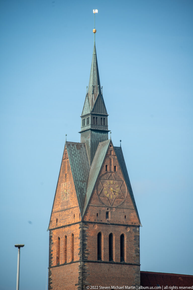 Brick church tower with green-patinated roof, louvered openings, and a large clock in a triangular gable.