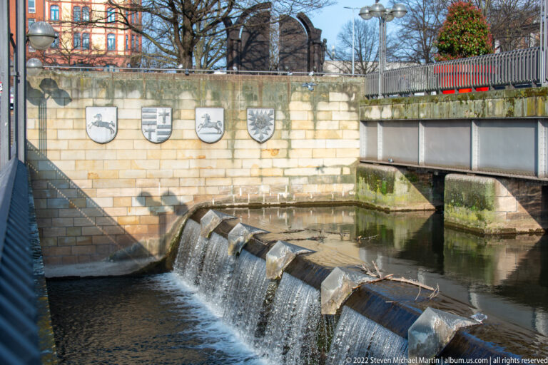 Low urban weir below a stone wall with heraldic shields and a bridge crossing the watercourse.
