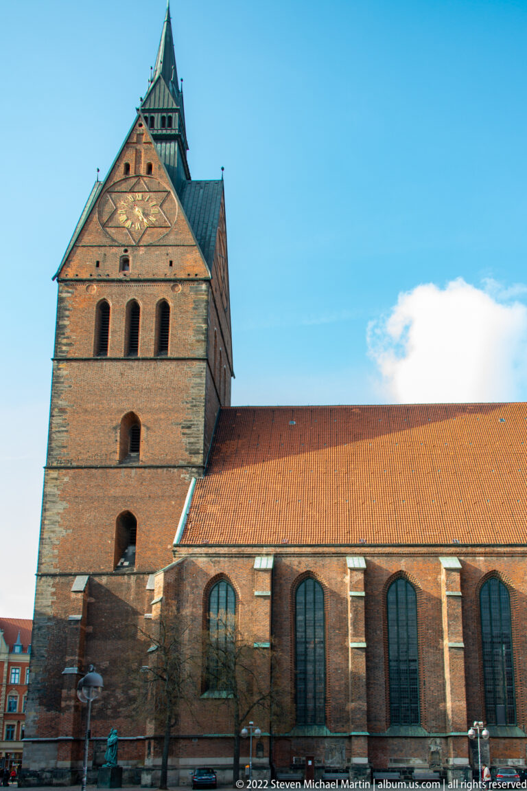 Brick church with massive square tower, long red roof, tall side windows, and pedestrians at street level.