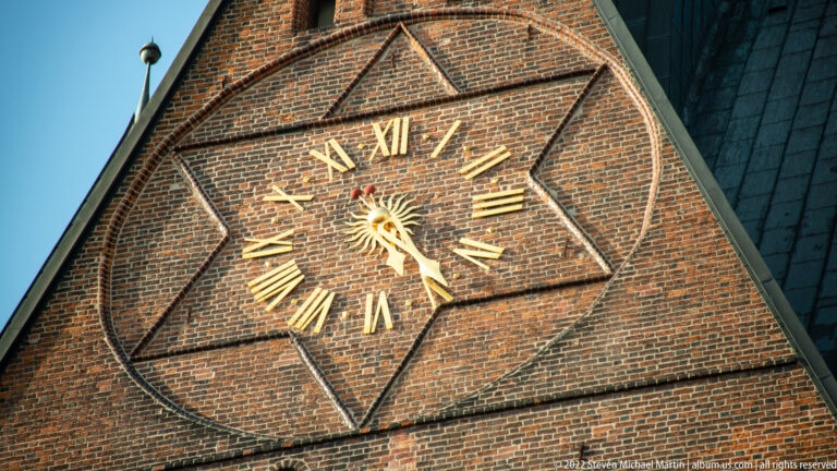 Large gilded Roman-numeral clock set into patterned red brick on a church gable.