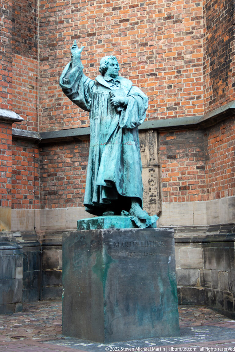 Green-patinated bronze statue of a standing man gesturing upward on a dark stone pedestal.