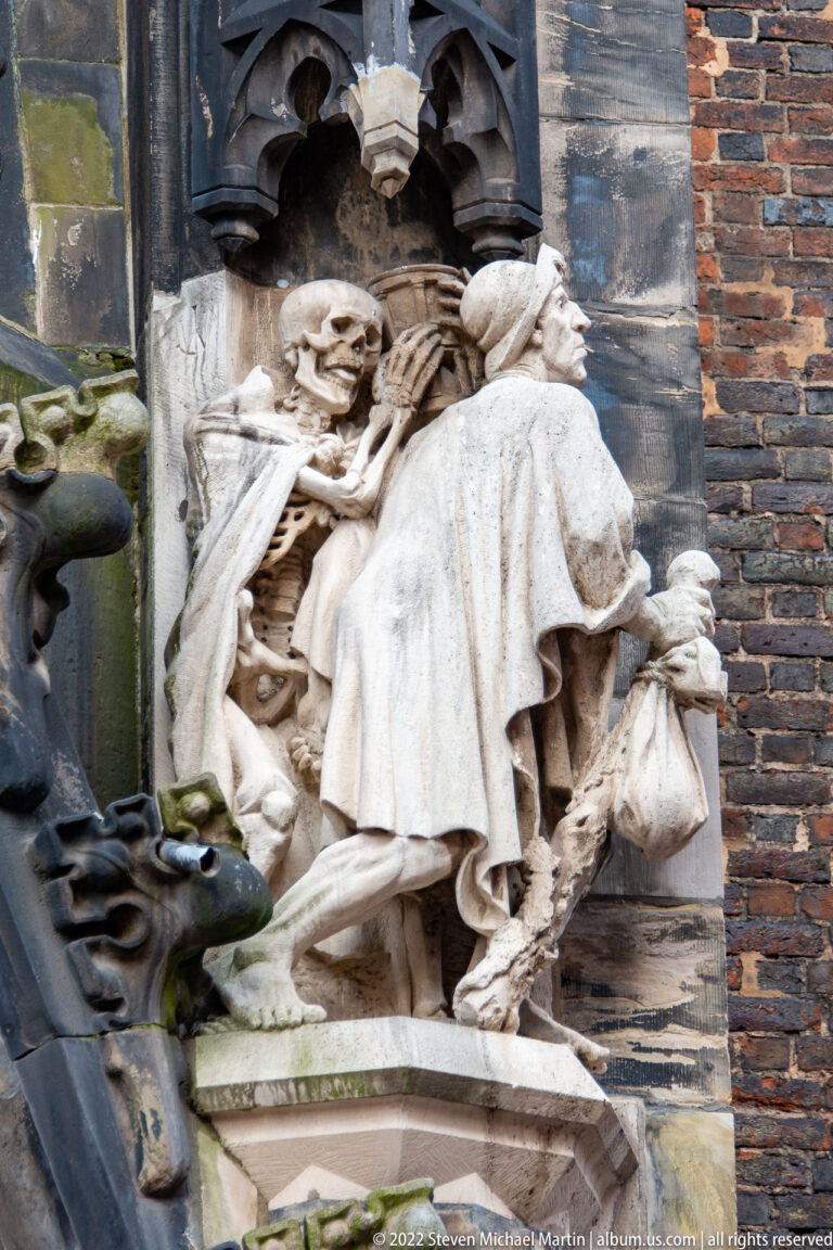 Weathered pale stone figure with deeply carved drapery set into a dark Gothic architectural surround.