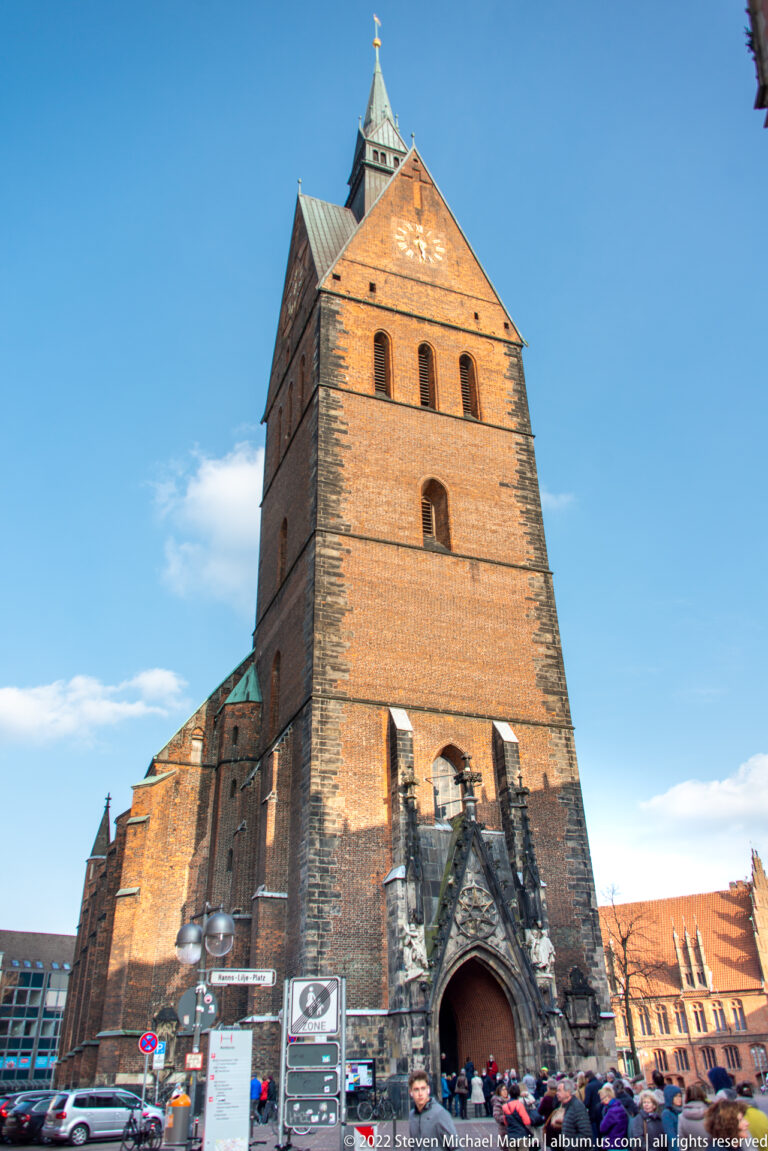 Massive brick church tower above a pointed portal, with pedestrians and signs at the base.
