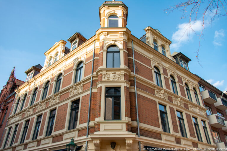 Ornate red-brick corner townhouse with pale stone trim, bay windows, dormers, and a varied roofline.
