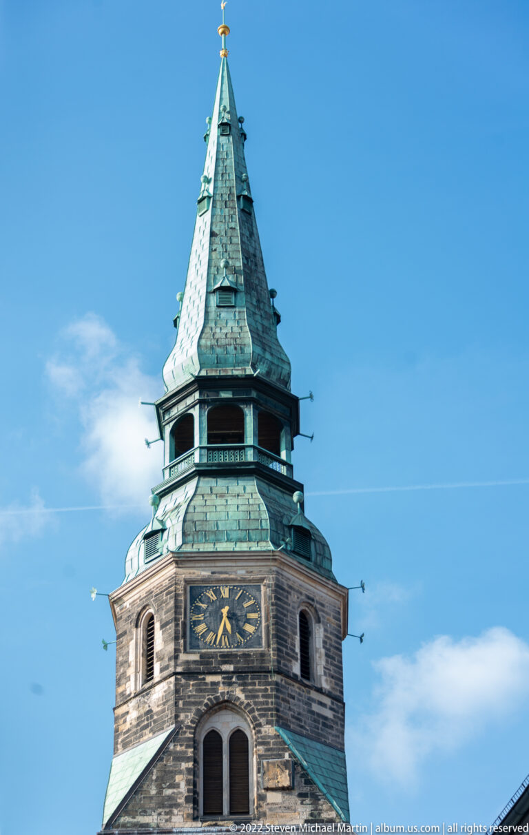 Upper church tower with arched openings, dark clock face, and steep green-patinated octagonal spire.